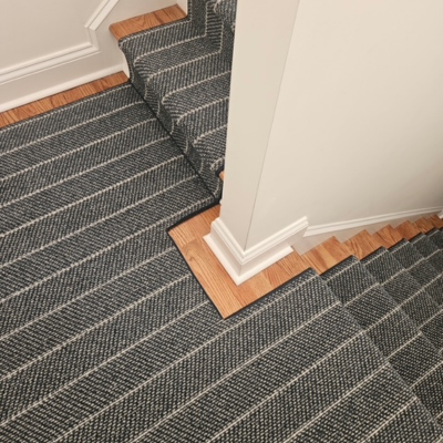 A staircase with a dark gray carpet runner featuring a herringbone pattern and white pinstripes, bordered by natural wood steps.